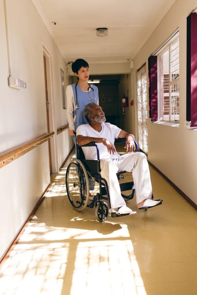 Female nurse standing while senior male sits in wheelchair in corridor at retirement home.